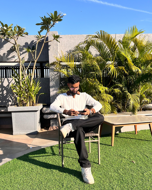 Person sitting outdoors on a sunny day wearing white polo and black linen pants with greenery and a clear blue sky.