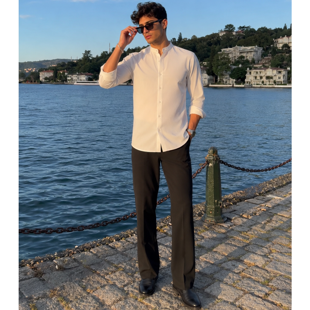 Man in white shirt and black pants standing by a waterfront with buildings and trees in the background.