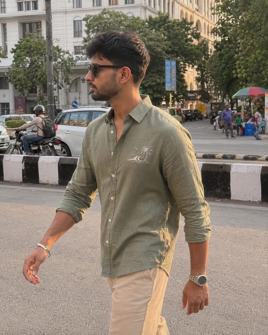 Man walking on a street wearing a green shirt with a logo, sunglasses, and a watch.