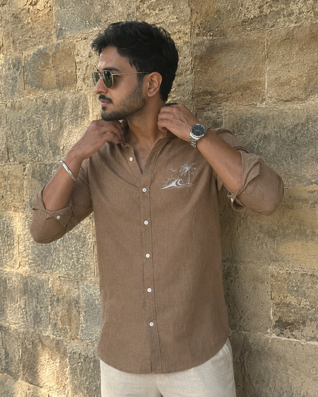 Man wearing a brown shirt and sunglasses against a stone wall