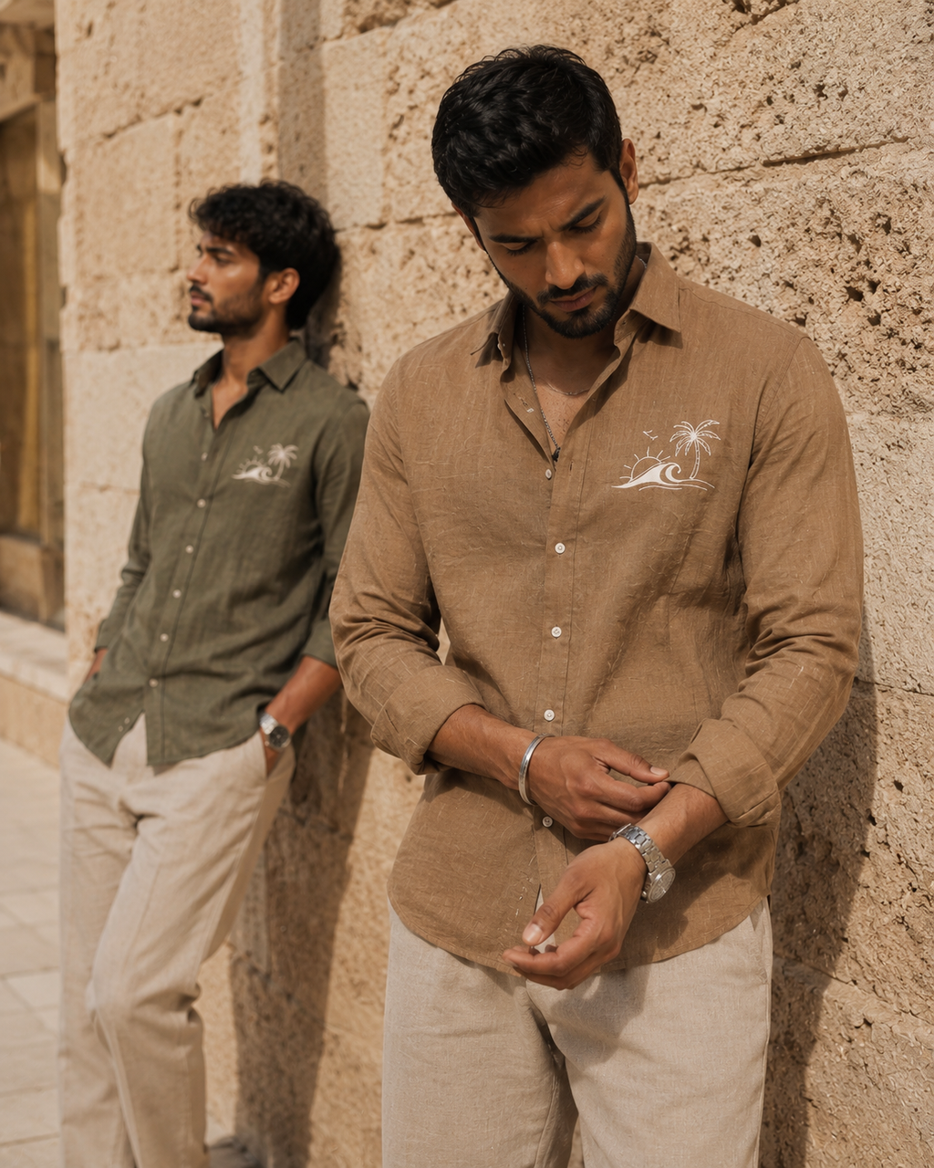 Two men wearing brown and green shirts with logos, standing against a stone wall.