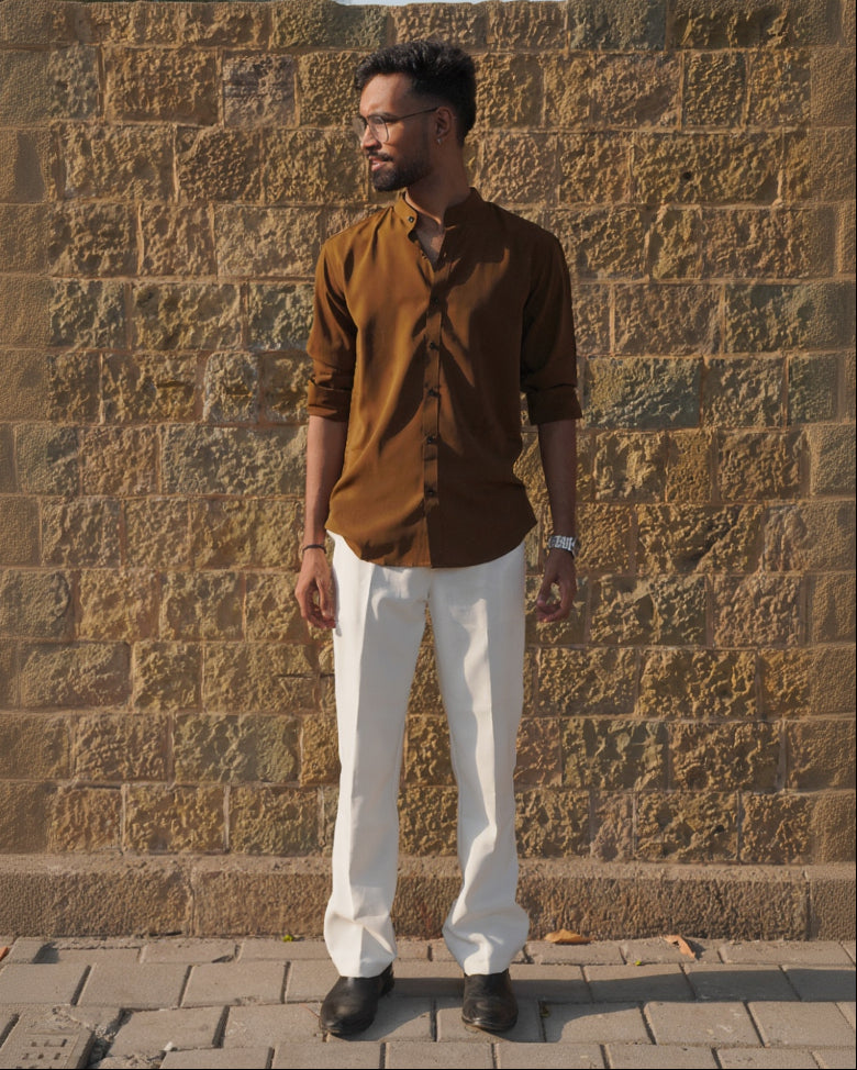 Man wearing a brown shirt and white pants standing against a stone wall.