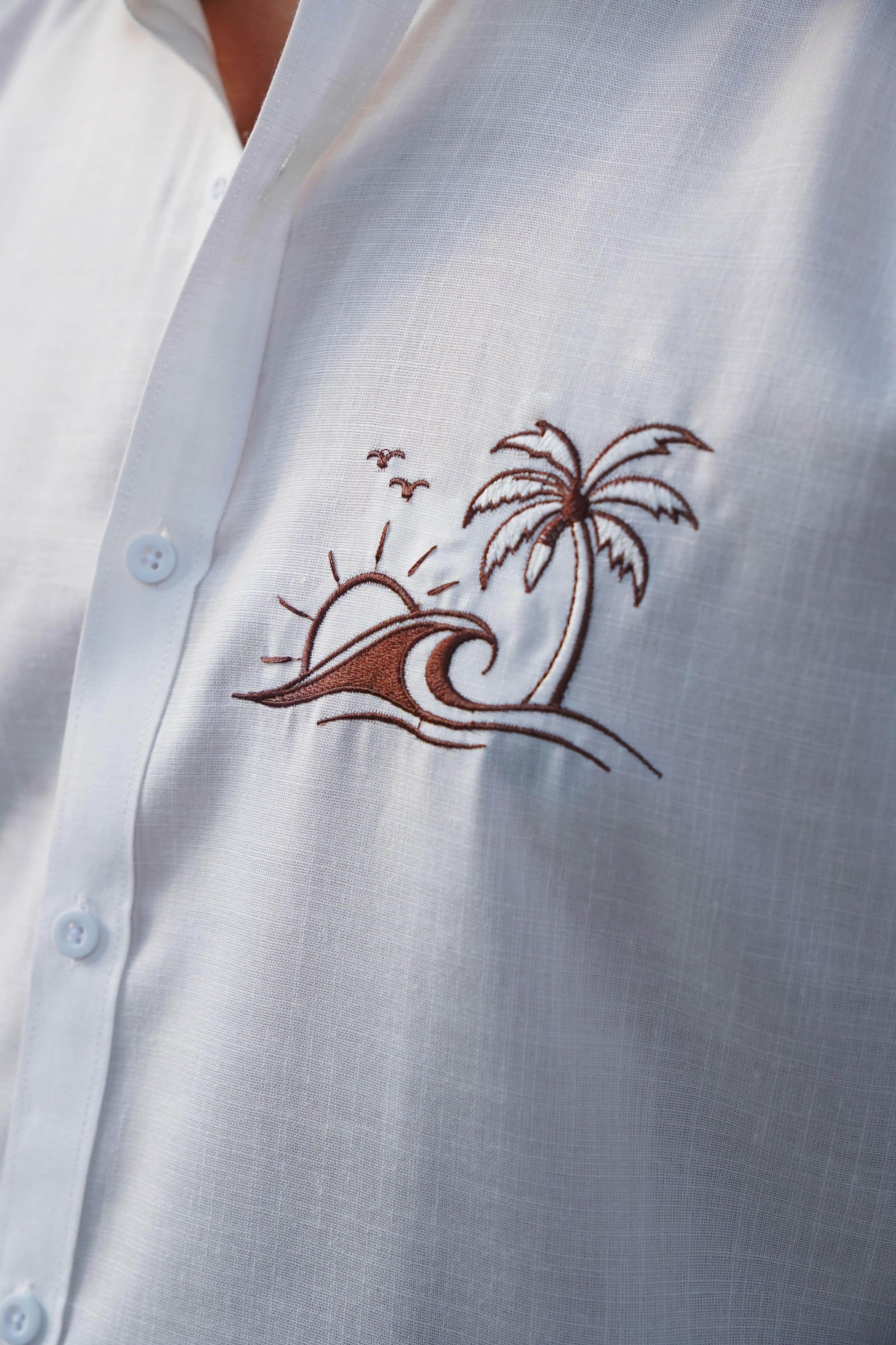 White shirt with brown embroidered palm tree and wave design.