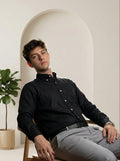 Man sitting on a chair wearing a black shirt and gray pants in a minimalistic setting.