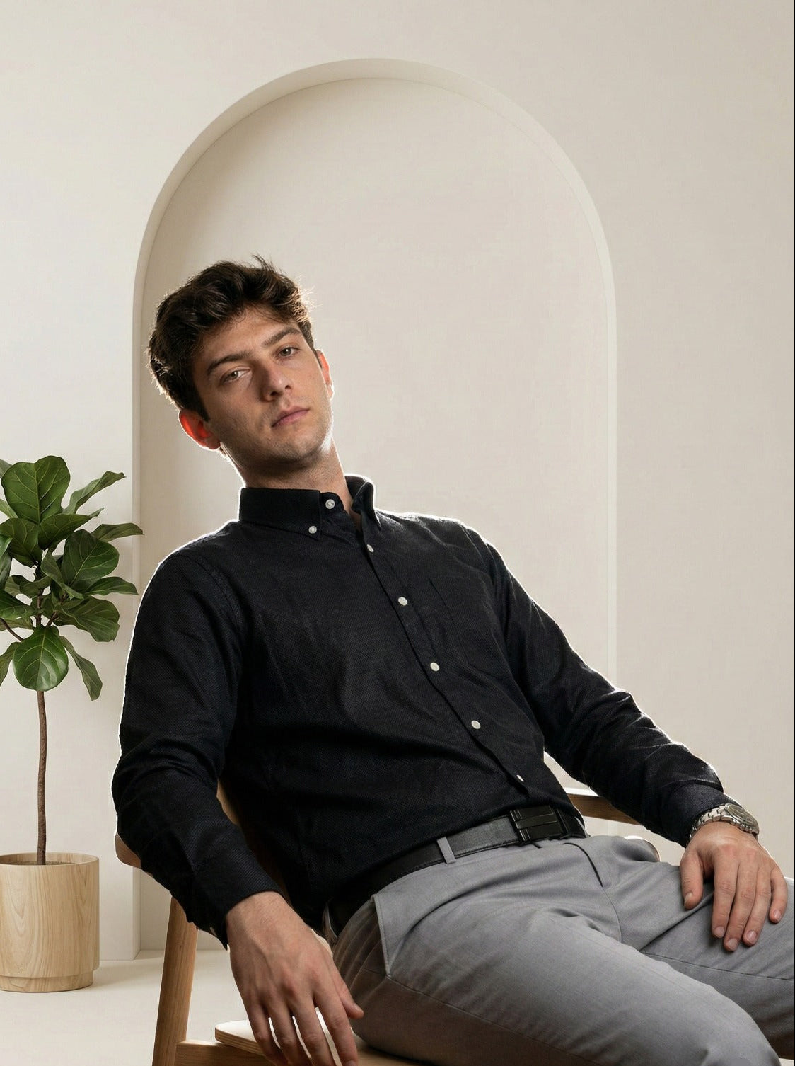 Man sitting on a chair wearing a black shirt and gray pants in a minimalistic setting.